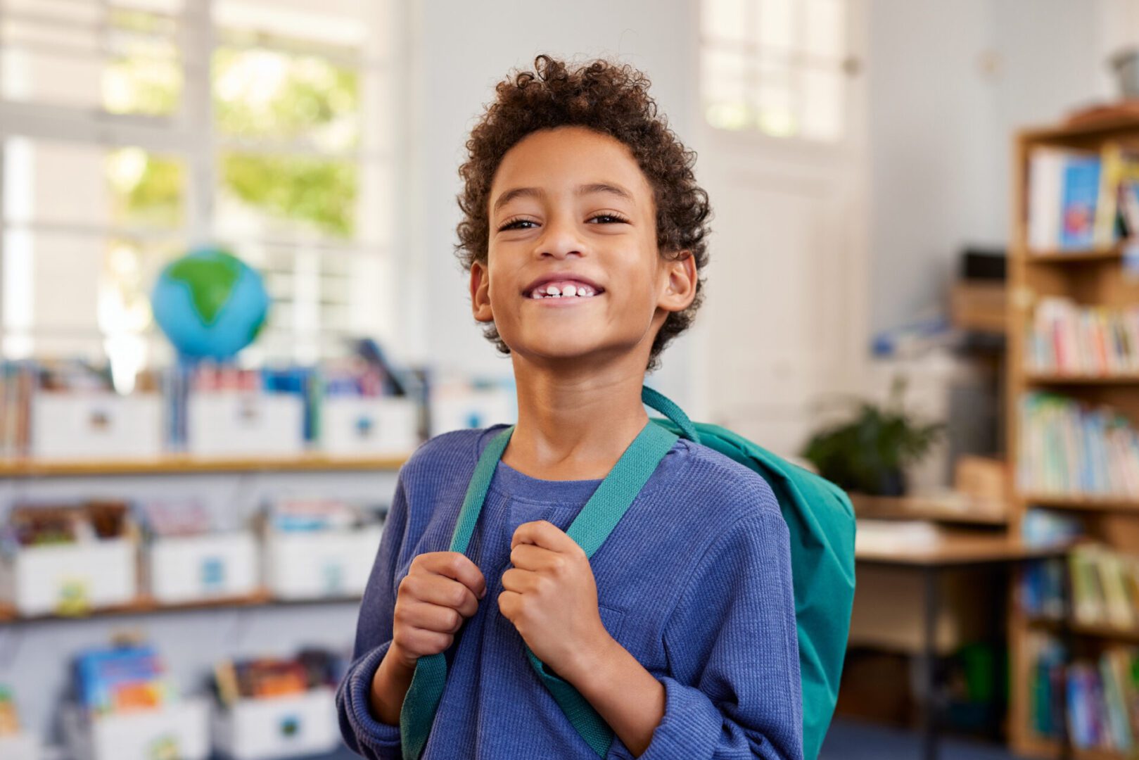 Portrait of smiling african american boy looking at camera while wearing green backpack. Black elementary schoolboy carrying backpack and standing in library at primary school. Cheerful mixed race child standing with library in background when returning to school after the holidays.